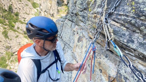 A man wearing a helmet and arrogance backpack looks at a pink and blue rope going through a set of chains connected to bolts installed in a rock face. 