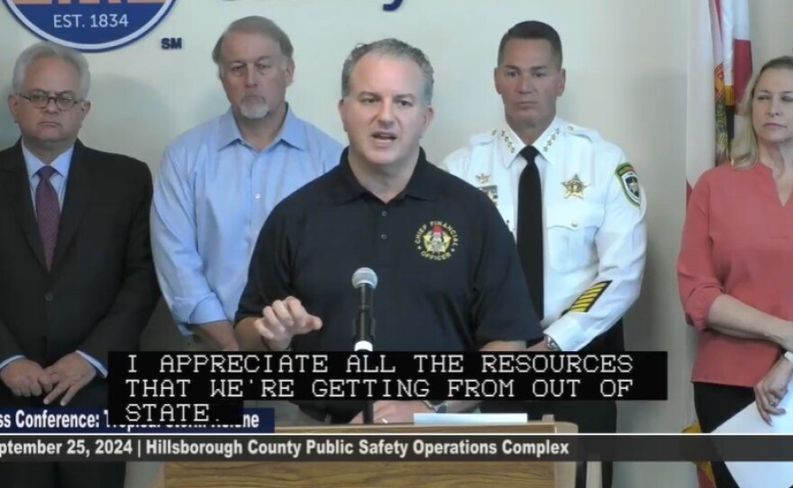 Man in a black shirt speaking at a podium with others in the background