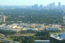 an aerial image showing a large interior shopping mall in the foregrown and downtown skyline through hazy sky toward the rear of the frame.  A major road on the right of the mall and image frame 
