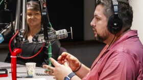 A man sitting in a radio studio gestures with both hand and appears to be talking at a microphone.