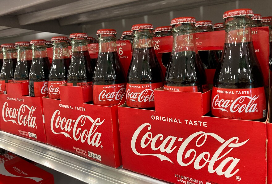 Glass bottles of Coca-Cola are stacked on a shelf in a grocery store.