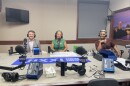 Three people wearing headphones sit at a table in a radio talk studio: a man at left has short brown hair, a brown beard and is wearing a light grey blazer over a white button-down shirt; a woman at center has shoulder-length grey hair and is wearing a green dress; a woman at right has short blonde hair and is wearing a brown dress.