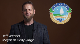 A screenshot from Holly Ridge mayor Jeff Wenzel's video announcement concerning Holly Plaza's rent vouchers. Mayor Wenzel is a white man with a brown beard and closely cropped brown hair. He's wearing a dark plaid sportcoat over a black t-shirt. He stands before a brown background, looking serious. On the right side of the image is the Holly Ridge town seal. The banner on the bottom reads "Jeff Wenzel, Mayor of Holly Ridge."