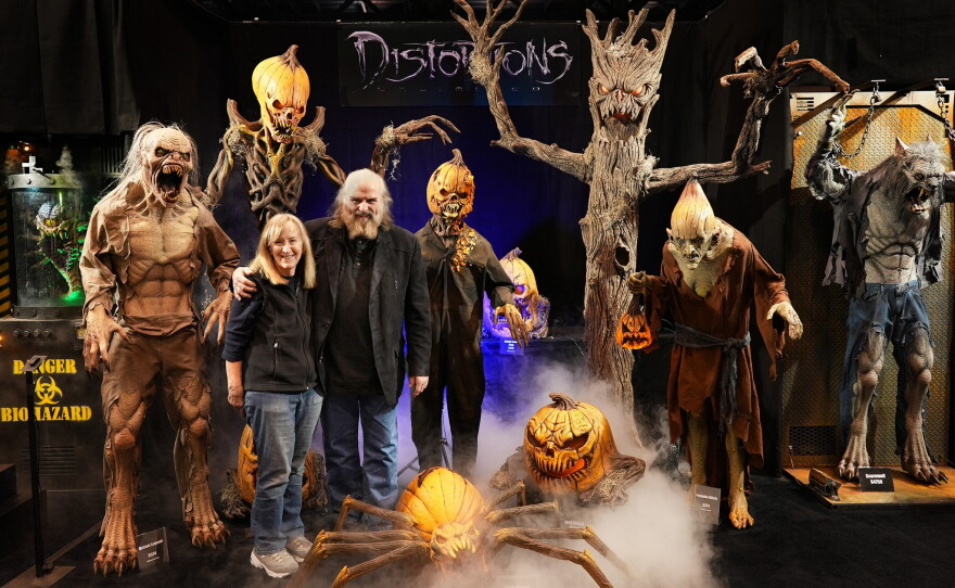 Marsha and Ed Edmunds stand amid life-size Halloween monsters including pumpkin-headed skeletons and a giant pumpkin spider