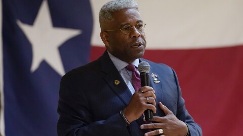 A man in a suit holds a microphone to his mouth in front of a Texas flag.