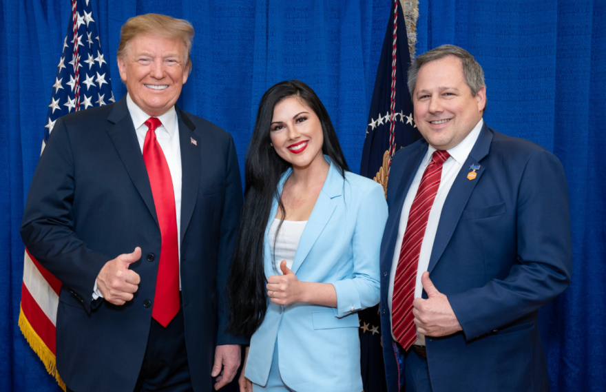 Photo shows three people who have one thumb up each. There is a taller man in a navy blue suit and red tie on the right. In the middle is a pretty brunette with long dark hair wearing a powder blue suit and a man on the right in a red, blue, and coral diagonal-striped tie. He is also wearing a blue suit. All three are smiling and look like they are enjoying themselves.