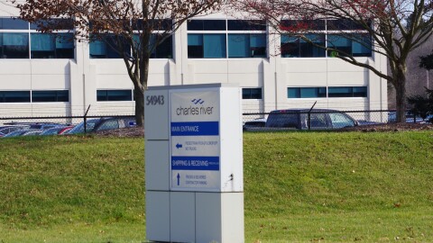 A white and blue sign advertises the entrance to the Charles River facility in Mattawan. It sits on the facilities deep green front lawn. Two trees sit behind it, atop the slight incline that separates the lawn from the facilities tall black gates topped with barbed wire. The bone white facility looms beyond the brief spattering of vehicles in the parking lot.  