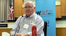 A man in a button-down shirt sits at a desk with a microphone pointed at him. 