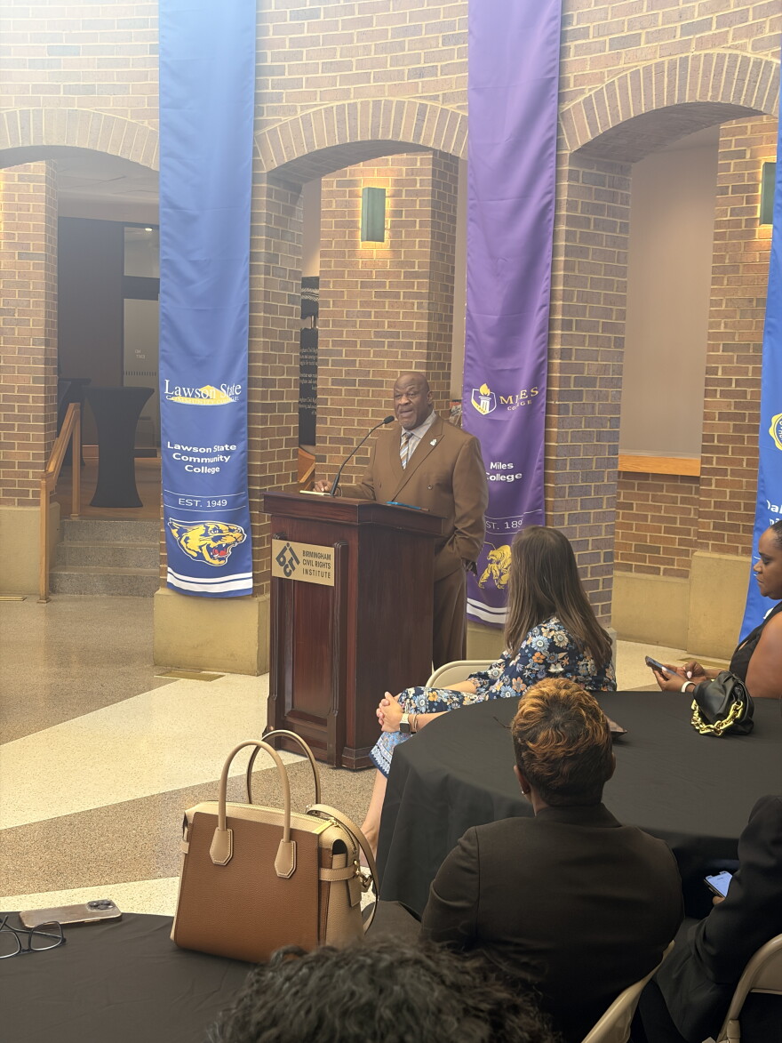 A man speaks behind a podium with three vertical banners behind him.