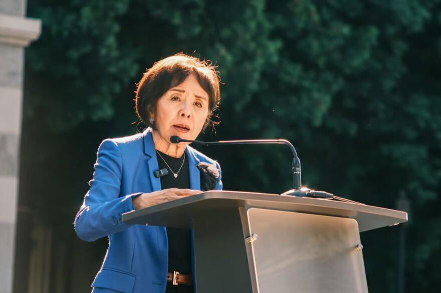 A woman wearing a blue blazer, with dark hair, speaks into a microphone on a lectern outdoors.