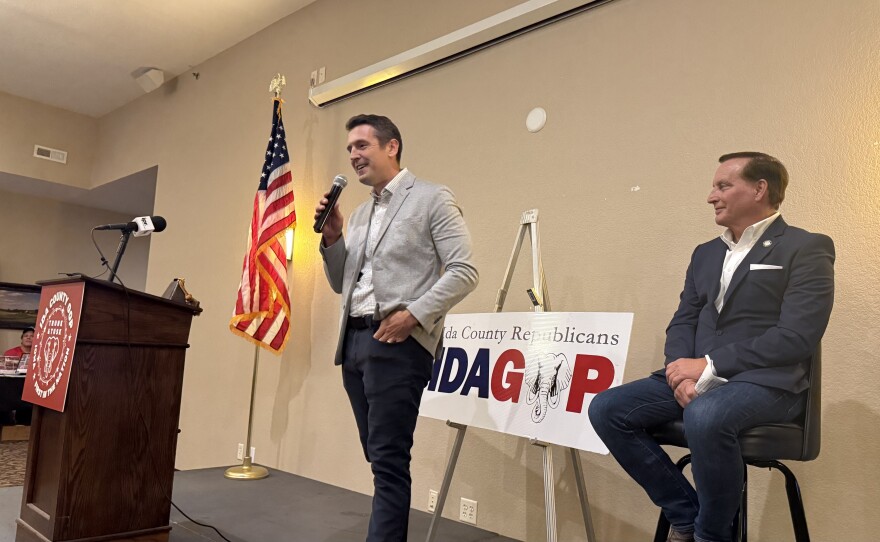 A man wearing a lighter colored jacket is holding a microphone. Another man is to the right.