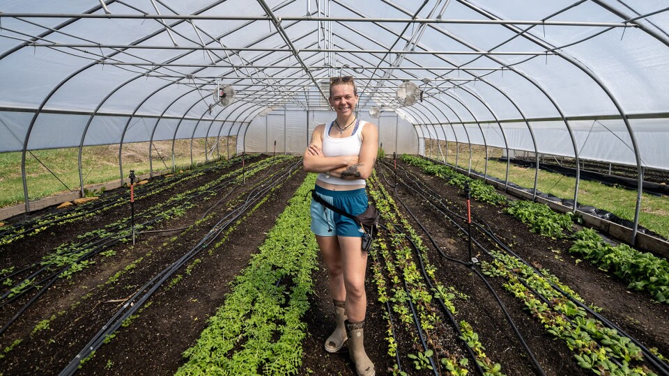 A person stands smiling with arms crossed inside a greenhouse, surrounded by rows of young plants on soil beds.