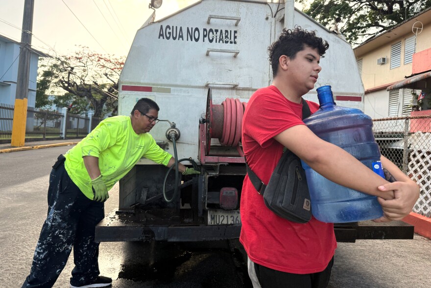 FILE - Bryan Pérez hauls a five-gallon water jug to his apartment
