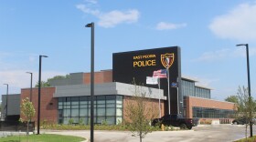 Exterior photo of the new East Peoria Police Department headquarters, a modern two-story building combining red brick, gray metal paneling, and a black accent wall with large illuminated "EAST PEORIA POLICE" lettering and the department's badge-shaped shield logo. Floor-to-ceiling glass windows wrap the lower level, with an American flag, a Thin Blue Line flag, and a city flag flying out front on tall poles.