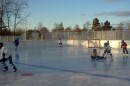 Youth hockey players are in various places on an outdoor hockey rink with a light blue sky and trees in the background.