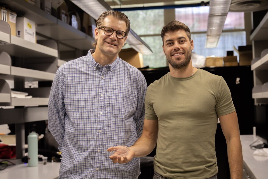 Two men stand in a laboratory holding a clear petri dish. 