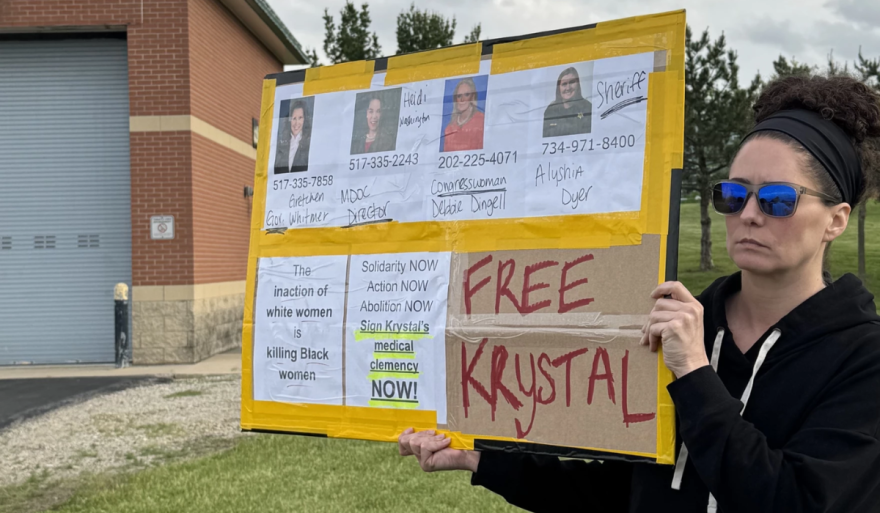 A person with sunglasses holds a yellow poster protesting for the release of an individual named Krystal. The sign displays contact numbers for Michigan officials, including Governor Gretchen Whitmer and Congresswoman Debbie Dingell. It also features phrases like "FREE KRYSTAL," "The inaction of white women is killing Black women," and "Sign Krystal's medical clemency NOW!"