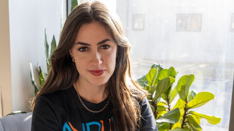 A young woman with long, brown hair gives the camera a half-smile. She's wearing an NPR t-shirt, a gold chain necklace and gold hoop earrings. Behind her are large-leafed plants and a sunlit (kind of dirt-streaked, if we're being honest) window.