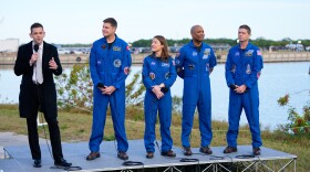 A man in a suit and holding a microphone speaks, while four people to the right wearing blue jumpsuits look at him. They're outdoors with a lake behind them.