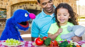 Blue Grover puppet from Sesame Street is with a young girl and her father. They are looking at healthy fruits and vegetables. 