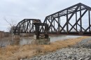 A rusted metal bridge spans a river. There are two concrete footings on the banks of the river that support the bridge.