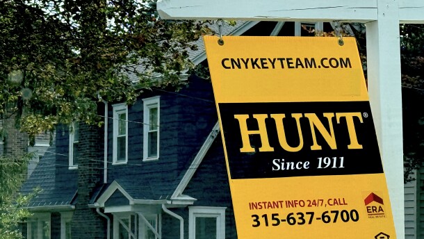 Yellow and black home sale sign swings on white post in front of a blue and white house.