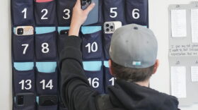 A ninth grader places his cellphone into a phone holder as he enters class at Delta High School, Friday, Feb. 23, 2024, in Delta, Utah. At the rural Utah school, there is a strict policy requiring students to check their phones at the door when entering every class. Each classroom has a cellphone storage unit that looks like an over-the-door shoe bag with three dozen smartphone-sized slots. 