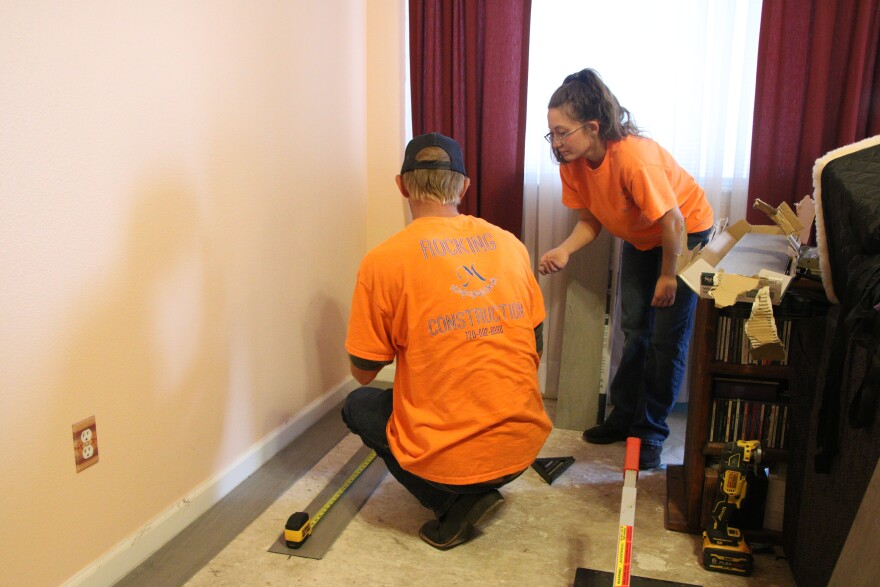 A man is crouching on the floor over some uninstalled grey floorboards. A woman is bending over him, talking with him about the project. There's some power tools on the floor.