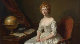 A fair-haired young woman in an ivory gown sits in a room with an old globe on the desk 
