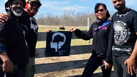 Four Black outdoor enthusiasts stand in a field beside a wooden fence.