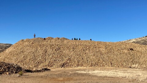 This is an image of a giant pile of mine waste, which looks like dirt and rock. People are standing on top of the brown-colored mound on a blue sky day.
