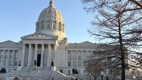 A photo of the exterior of the Missouri State capitol building. Vehicles are parked in front of the domed structure, people walk on the sidewalk. 