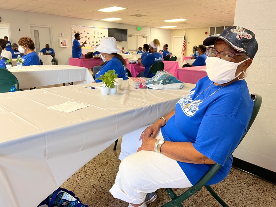 Woman sits wearing a face mask. Tables at a senior center are spread out around the room with other elderly people wearing masks sitting at them.