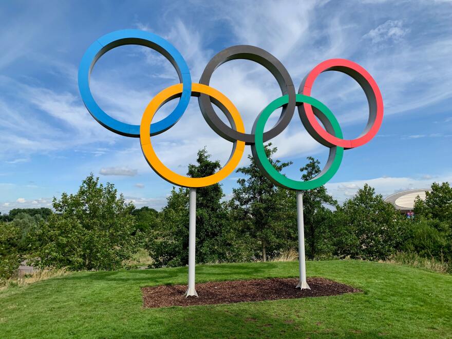 The blue, yellow, black, green, and red Olympic rings sit suspended in the air against a blue sky.