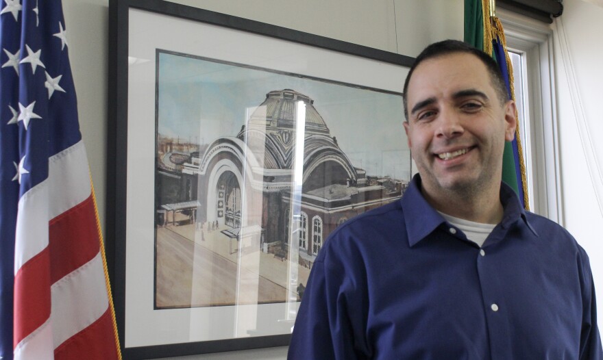Tacoma Mayor Anders Ibsen stands in front of a painting and alongside the U.S. and Washington flags.