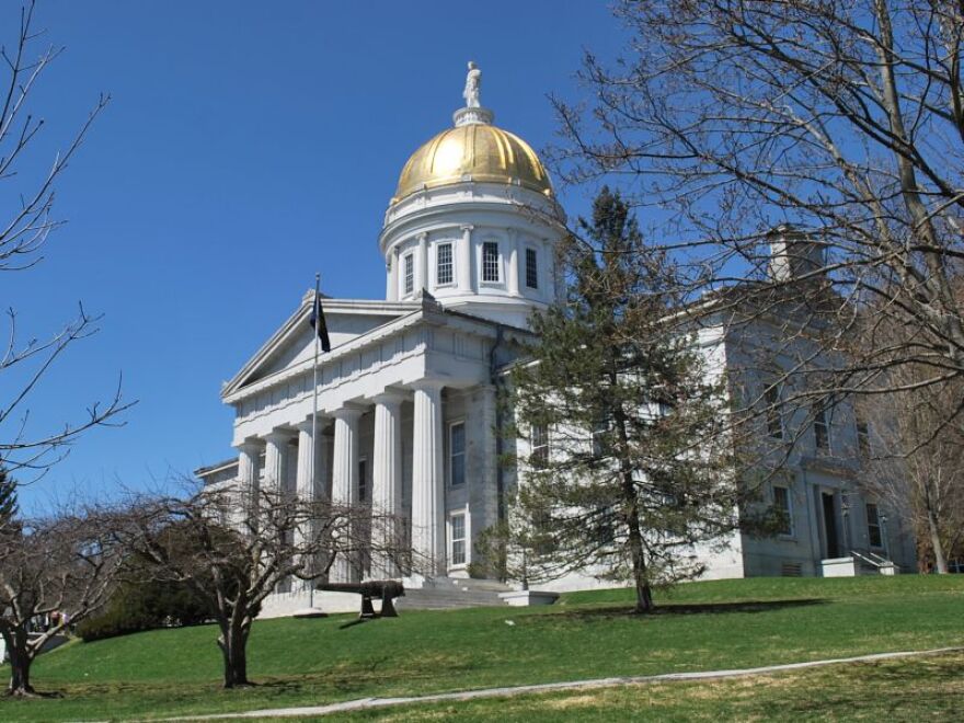 The Vermont Statehouse, a building with white columns and a giant golden dome, stands beyond a field of grass and trees.