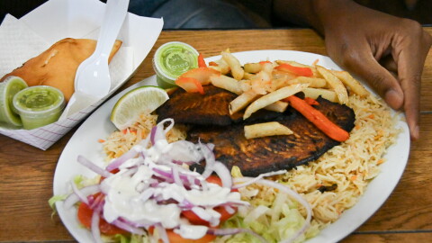 An oval white plate sits on a table. A man's hand is touching the side. The plate shows a salad with white dressing, rice and blackened fish. Beside the plate is a cardboard container with a brown, fried pastry.