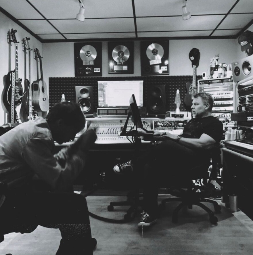 Two people are in a music studio with guitars on the wall, sound equipment, and framed awards. One person sits by a mixing console, and the other is in the foreground, head down. The image is in black and white.