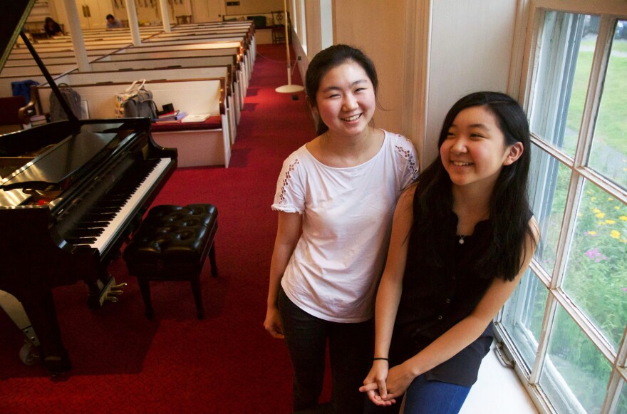 Two girls stand by a piano.