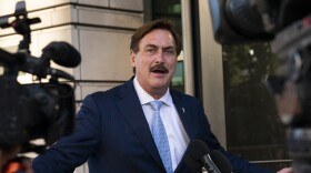 A white man in a navy blue suit with a white button-down shirt and light blue tie. He has brown hair and a brown mustache.