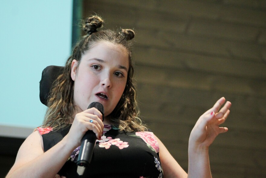 Kendra Hummer speaks into a handheld microphone at a community town hall. She is a white woman who uses a motorized wheelchair as a mobility aid. She wears a black dress with a pink flower print on it. She has brown shoulder-length hair and about half of it is in two small space buns on the top of her head. She holds the microphone in her right hand and gestures with her left hand. Behind her is the corner of a presentation screen which has a simple white slide on it. 