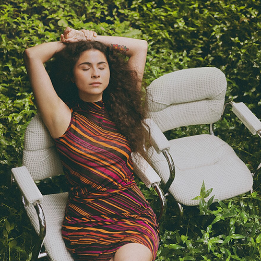 A woman sits in an white chair with her arms over her head. Behind her are lush green leaves.