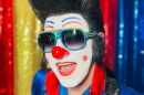 A man with clown face make up also looks like Elvis Presely with a fake black updo wig and a red nose. He is smiling.