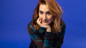 Sam Maggs sits in front of a royal blue background. She wears white tennis shoes and a patterned jackets. She has medium length red hair and she is sitting crosslegged with her head in her palm.
