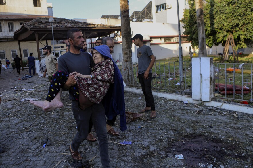 Wed., Oct. 18: A Palestinian man carries an elderly woman past the site of a deadly explosion at al-Ahli hospital, in Gaza City.