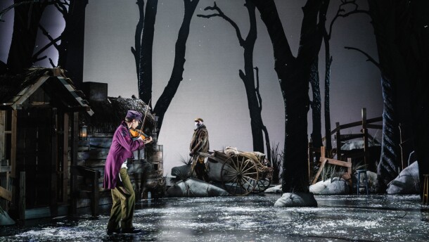 A young man wearing a purple coat and cap plays the violin on stage. In the background, an older man, wearing a villager costume, gazes at the violonist, while pulling a cart.