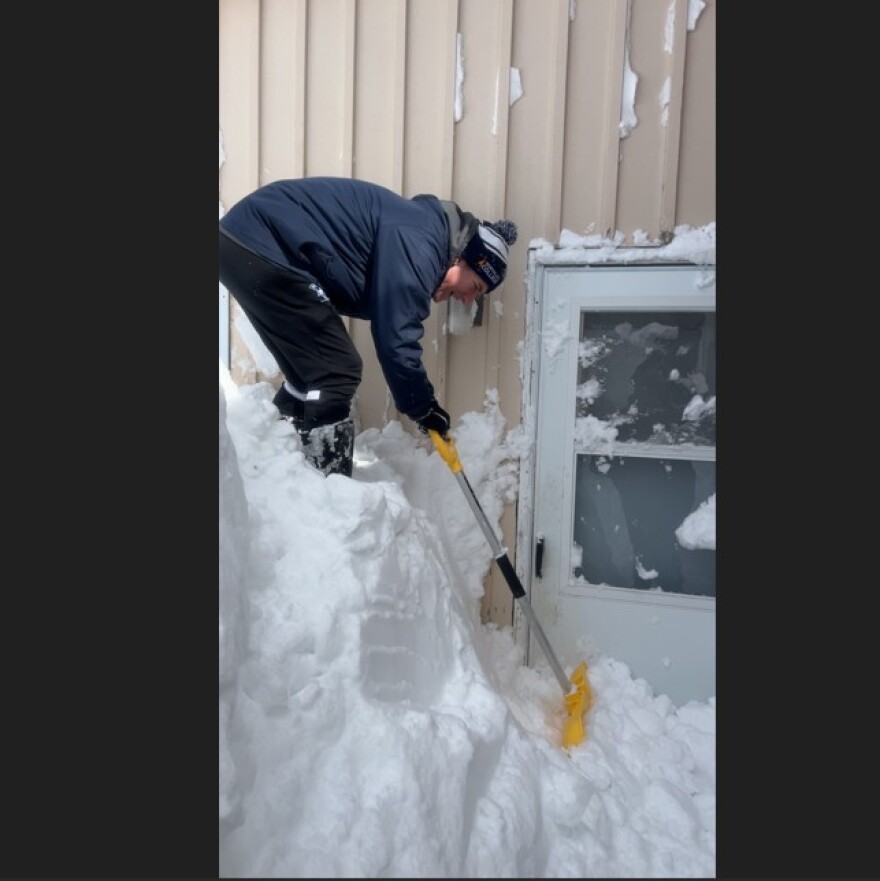 Street outreach team worker Chelan Gonzalez stands on top of a huge snowdrift, digging from the top down to clear the door to one of the family shelters run by Room at the Inn, a homeless shelter in Marquette, in March, 2026.