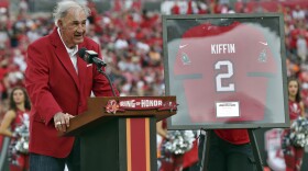 Monte Kiffin wearing a red sportcoat standing at a podium that says Ring of Honor and a framed Kiffin 2 jersey in glass next to him