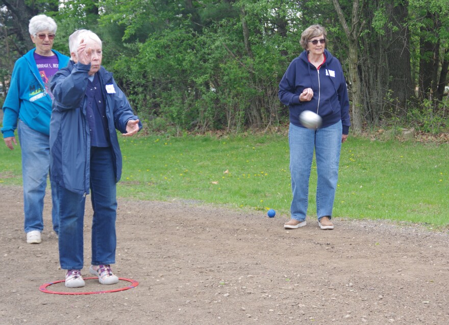 Woman in blue jacket and jeans throws petanque ball which can be seen as a blur near the right third of the frame, while two other women look on