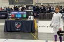 Jubilee holds a fencing helmet in her right hand and a fencing sword in her left hand. He also wears a white uniform. 